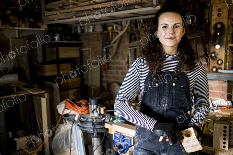Woman with long brown hair wearing dungarees standing in wood workshop, smiling at camera.