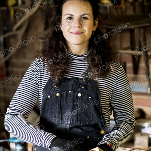 Woman with long brown hair wearing dungarees standing in wood workshop, smiling at camera. Woman with long brown hair wearing dungarees standing in wood workshop, smiling at camera.