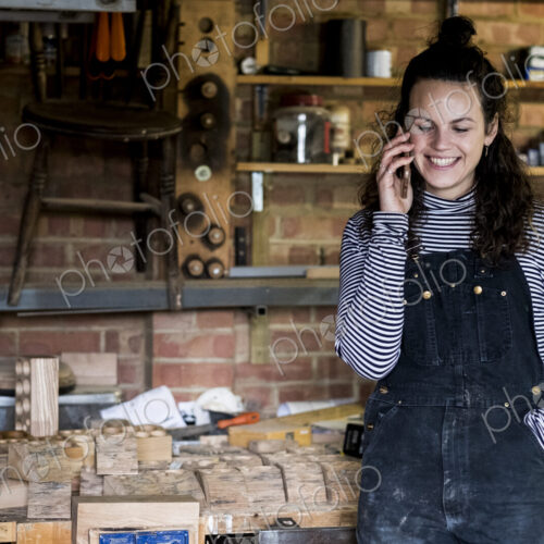 Woman with long brown hair wearing dungarees standing in wood workshop, talking on… Woman with long brown hair wearing dungarees standing in wood workshop, talking on…