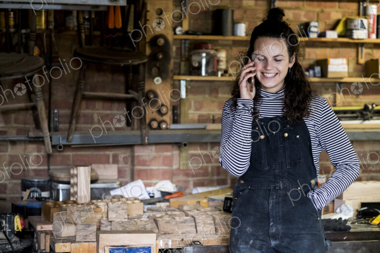 Woman with long brown hair wearing dungarees standing in wood workshop, talking on…