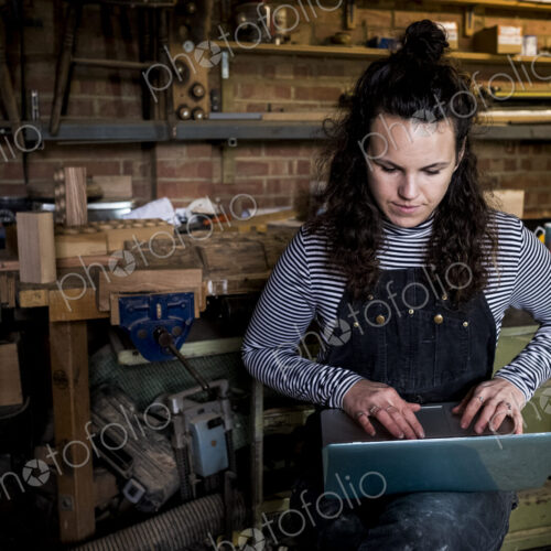 Woman with long brown hair wearing dungarees standing in wood workshop, typing on… Woman with long brown hair wearing dungarees standing in wood workshop, typing on…