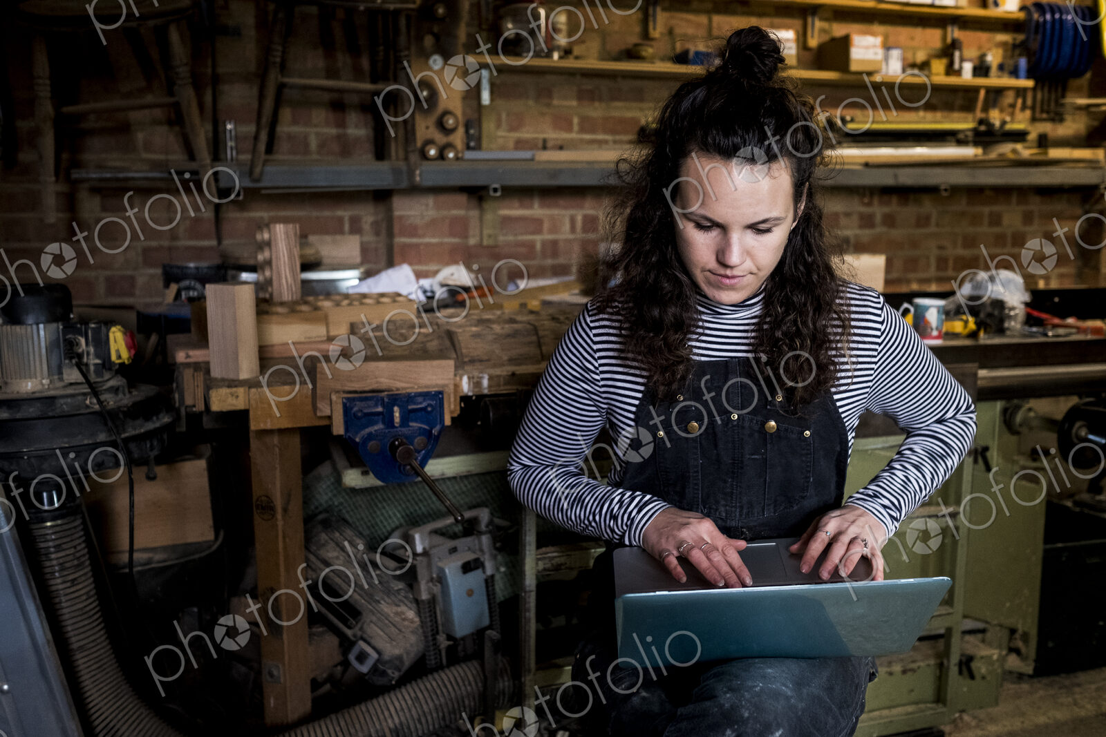 Woman with long brown hair wearing dungarees standing in wood workshop, typing on…