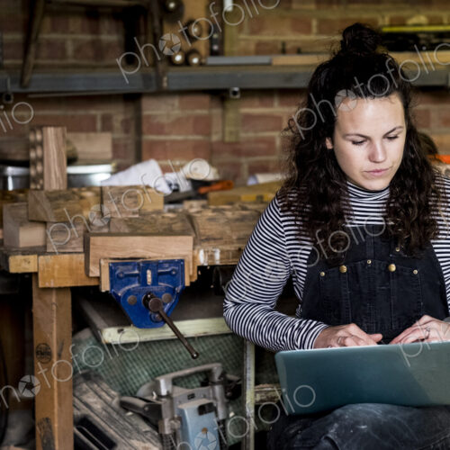 Woman with long brown hair wearing dungarees standing in wood workshop, typing on… Woman with long brown hair wearing dungarees standing in wood workshop, typing on…