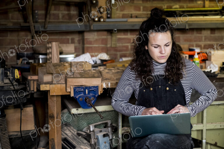 Woman with long brown hair wearing dungarees standing in wood workshop, typing on…