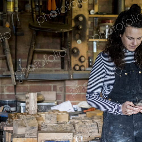 Woman with long brown hair wearing dungarees standing in wood workshop, using mobile… Woman with long brown hair wearing dungarees standing in wood workshop, using mobile…
