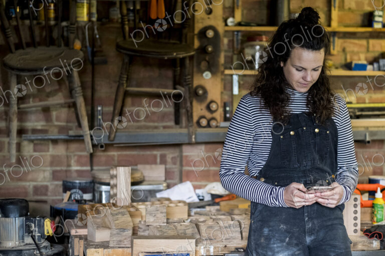Woman with long brown hair wearing dungarees standing in wood workshop, using mobile…