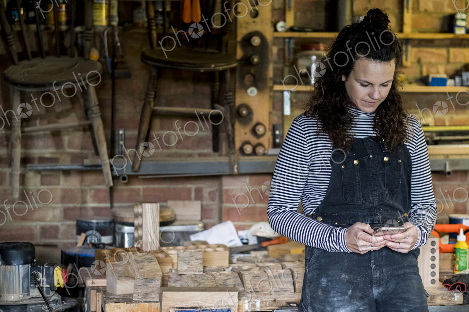 Woman with long brown hair wearing dungarees standing in wood workshop, using mobile…