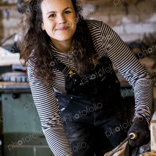 Woman with long brown hair wearing dungarees standing in wood workshop, using plane on… Woman with long brown hair wearing dungarees standing in wood workshop, using plane on…