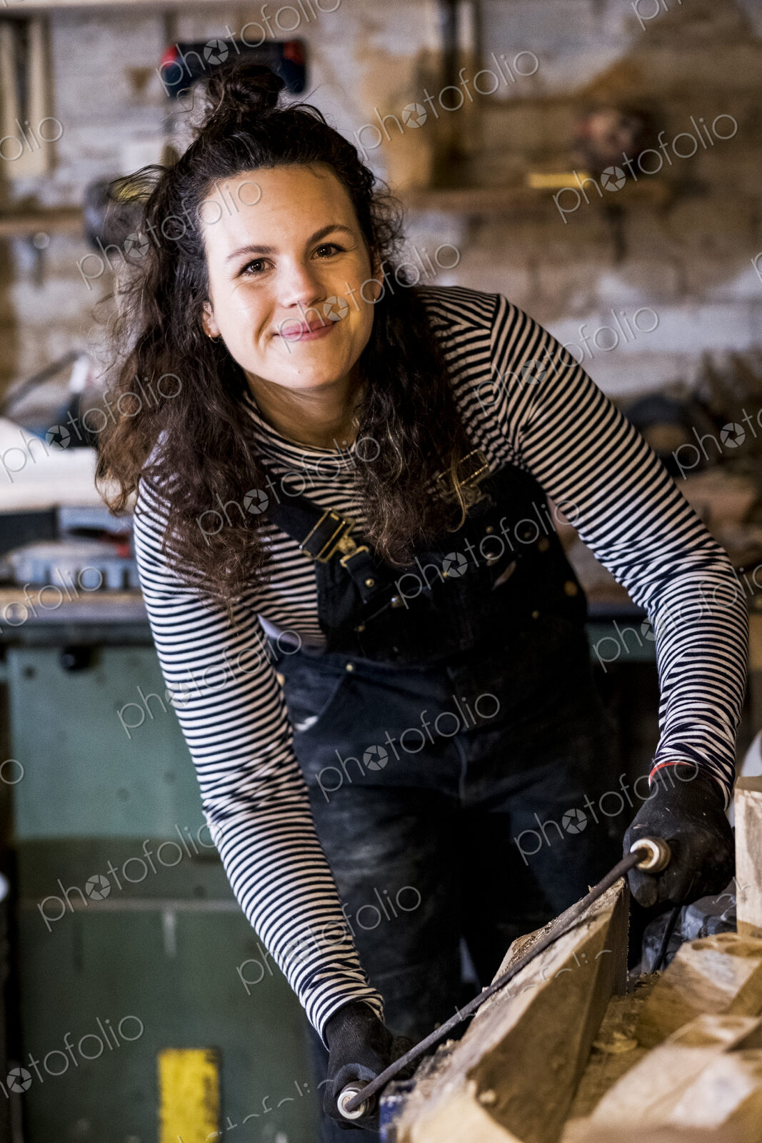Woman with long brown hair wearing dungarees standing in wood workshop, using plane on…