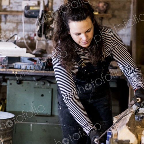 Woman with long brown hair wearing dungarees standing in wood workshop, using plane on… Woman with long brown hair wearing dungarees standing in wood workshop, using plane on…