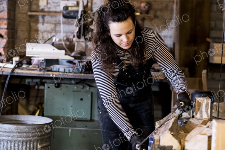 Woman with long brown hair wearing dungarees standing in wood workshop, using plane on…