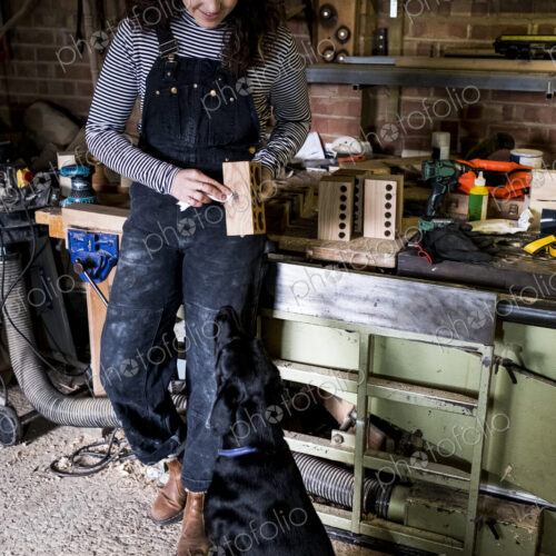 Woman with long brown hair wearing dungarees standing in wood workshop with black…
