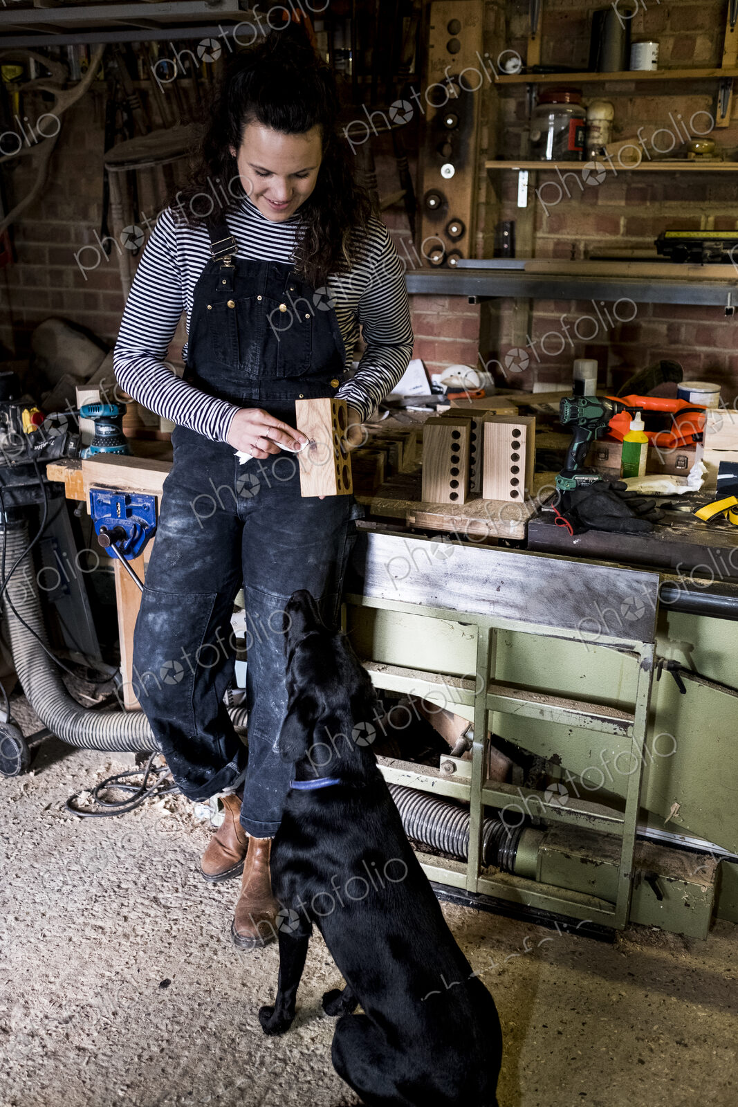 Woman with long brown hair wearing dungarees standing in wood workshop with black…