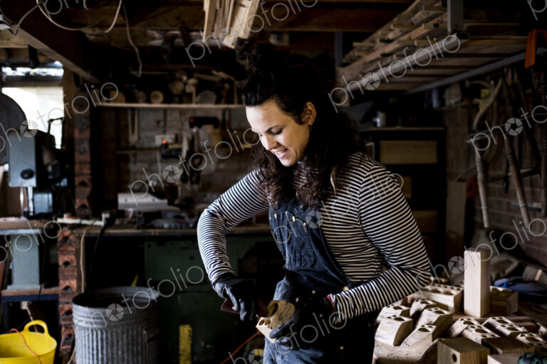 Woman with long brown hair wearing dungarees, working in wood workshop.