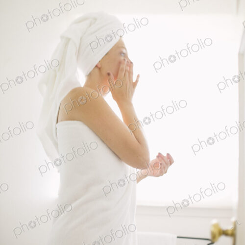 Woman wrapped in a white towel standing in a bathroom, applying cream to her face. Woman wrapped in a white towel standing in a bathroom, applying cream to her face.