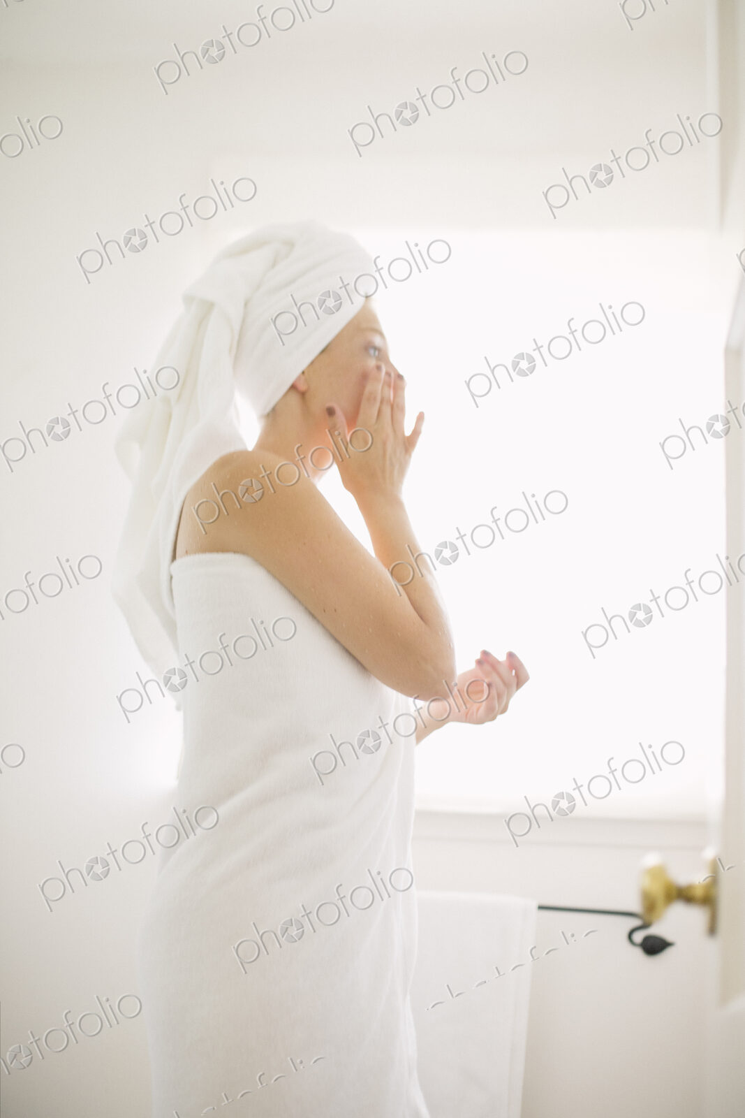 Woman wrapped in a white towel standing in a bathroom, applying cream to her face.
