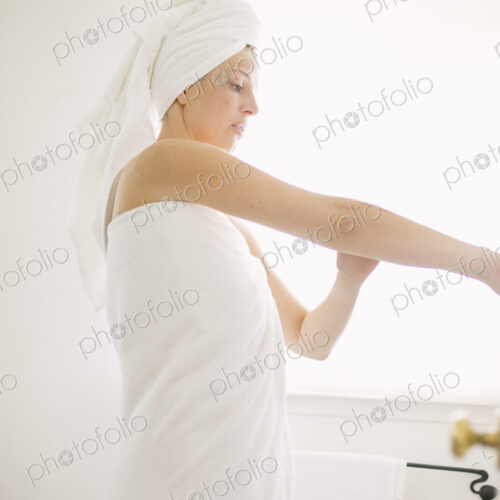 Woman wrapped in a white towel standing in a bathroom, applying lotion to her arm. Woman wrapped in a white towel standing in a bathroom, applying lotion to her arm.
