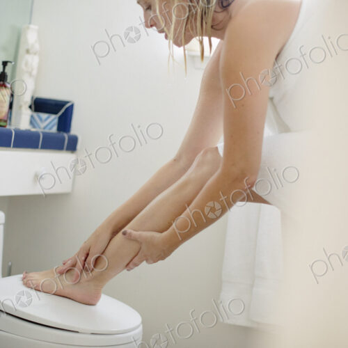 Woman wrapped in a white towel standing in a bathroom, applying lotion to her leg.