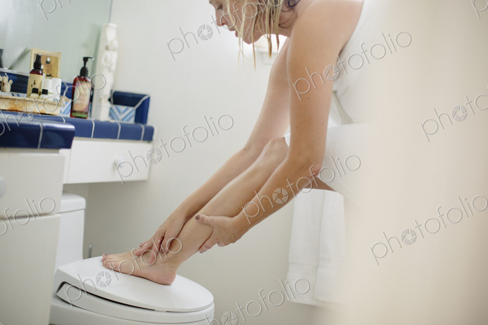 Woman wrapped in a white towel standing in a bathroom, applying lotion to her leg.