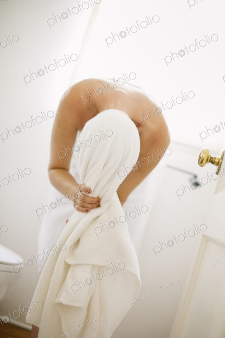 Woman wrapped in a white towel standing in a bathroom, wrapping her hair in a towel.
