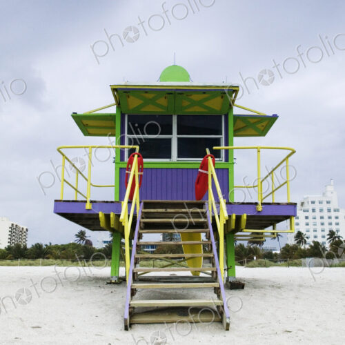 Wooden colourful lifeguard hut on a sandy beach with high rise buildings beyond.