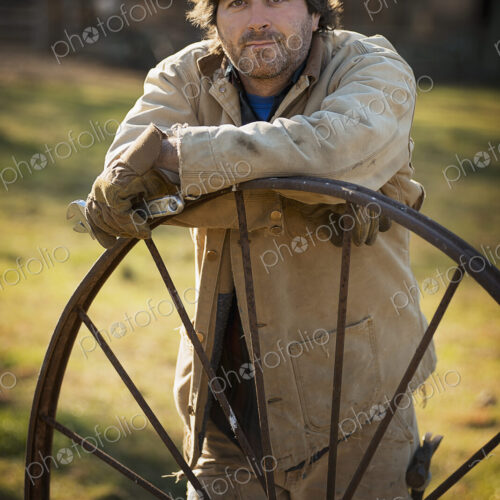 Working on an organic farm. A man in overalls wearing work gloves, leaning on a round… Working on an organic farm. A man in overalls wearing work gloves, leaning on a round…