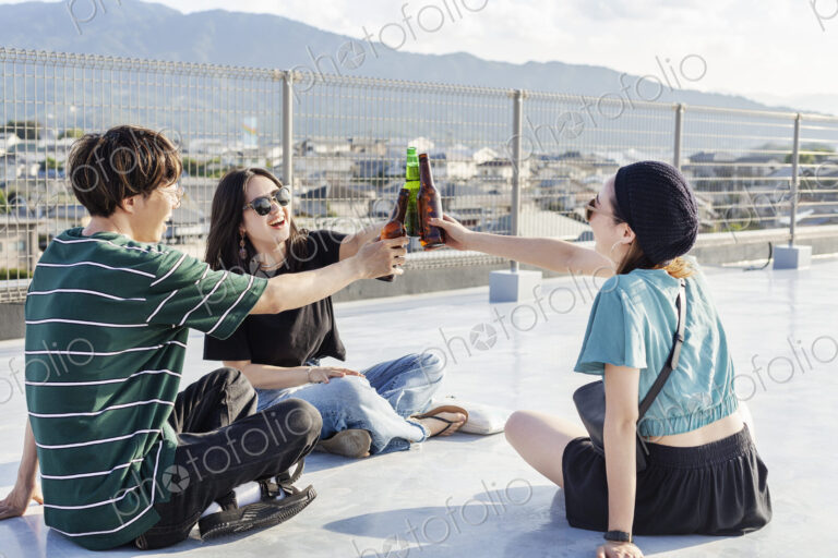 Young Japanese man and two women sitting on a rooftop in an urban setting, drinking beer.