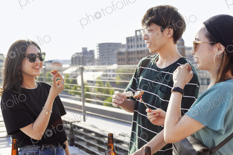 Young Japanese man and two women standing on a rooftop in an urban setting, drinking beer.