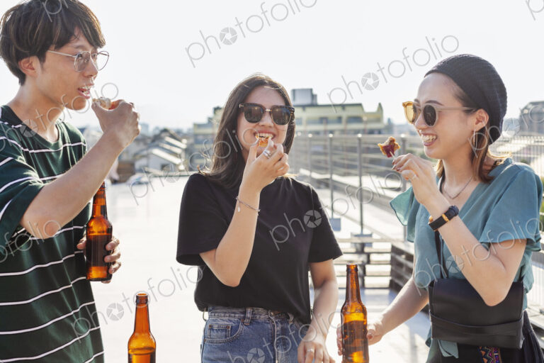 Young Japanese man and two women standing on a rooftop in an urban setting, drinking beer.