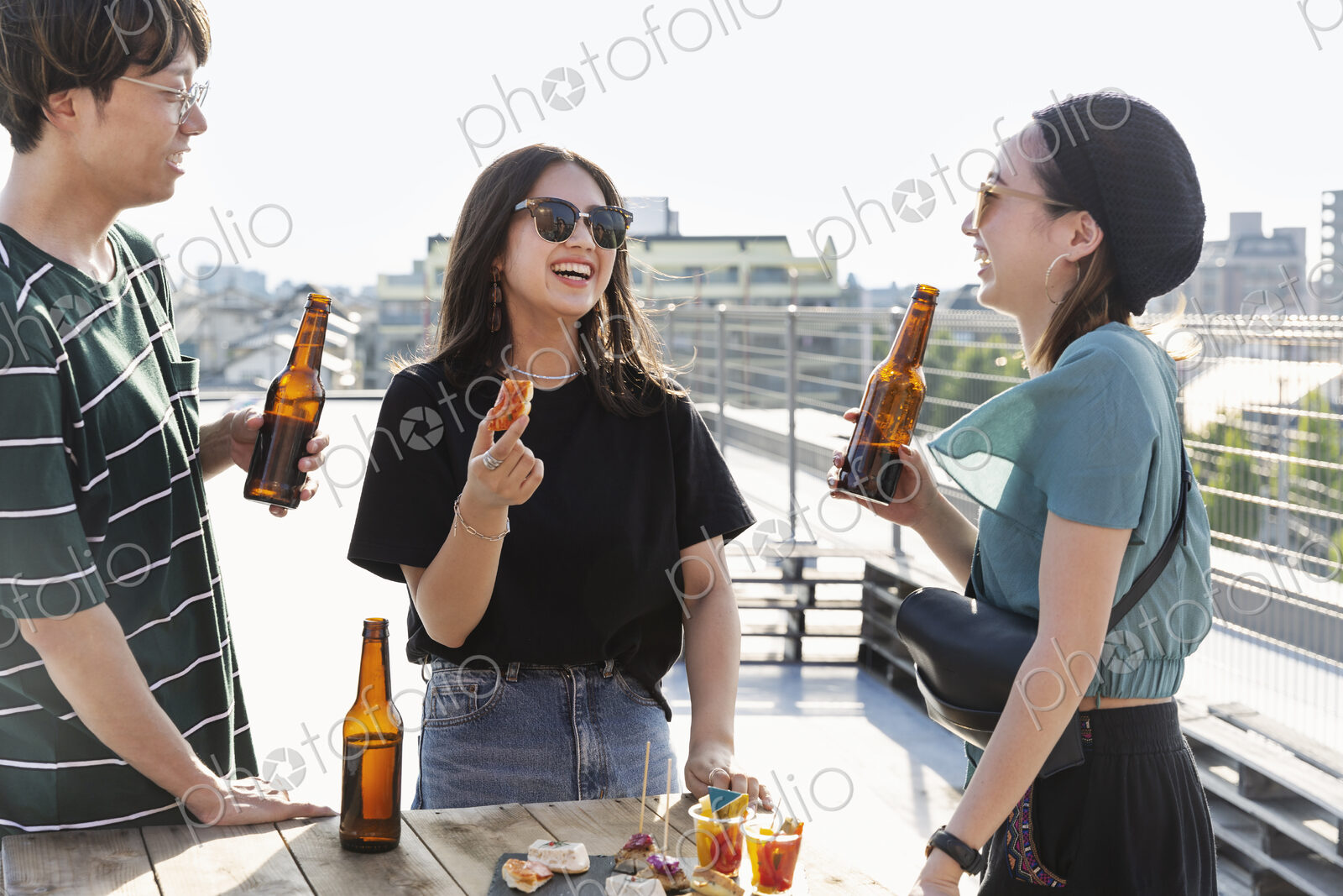 Young Japanese man and two women standing on a rooftop in an urban setting, drinking beer.