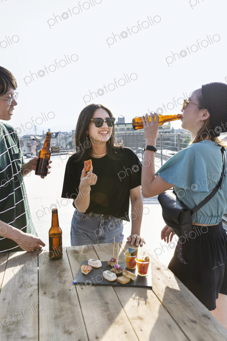 Young Japanese man and two women standing on a rooftop in an urban setting, drinking beer.