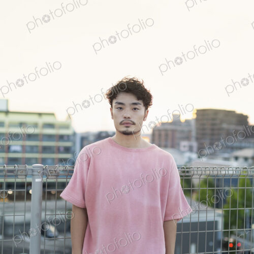 Young Japanese man standing on a rooftop in an urban setting, looking at camera.