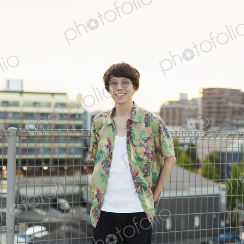 Young Japanese man standing on a rooftop in an urban setting, looking at camera.