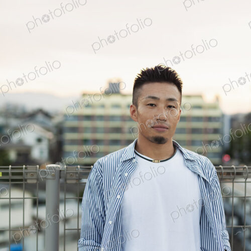 Young Japanese man standing on a rooftop in an urban setting, looking at camera.