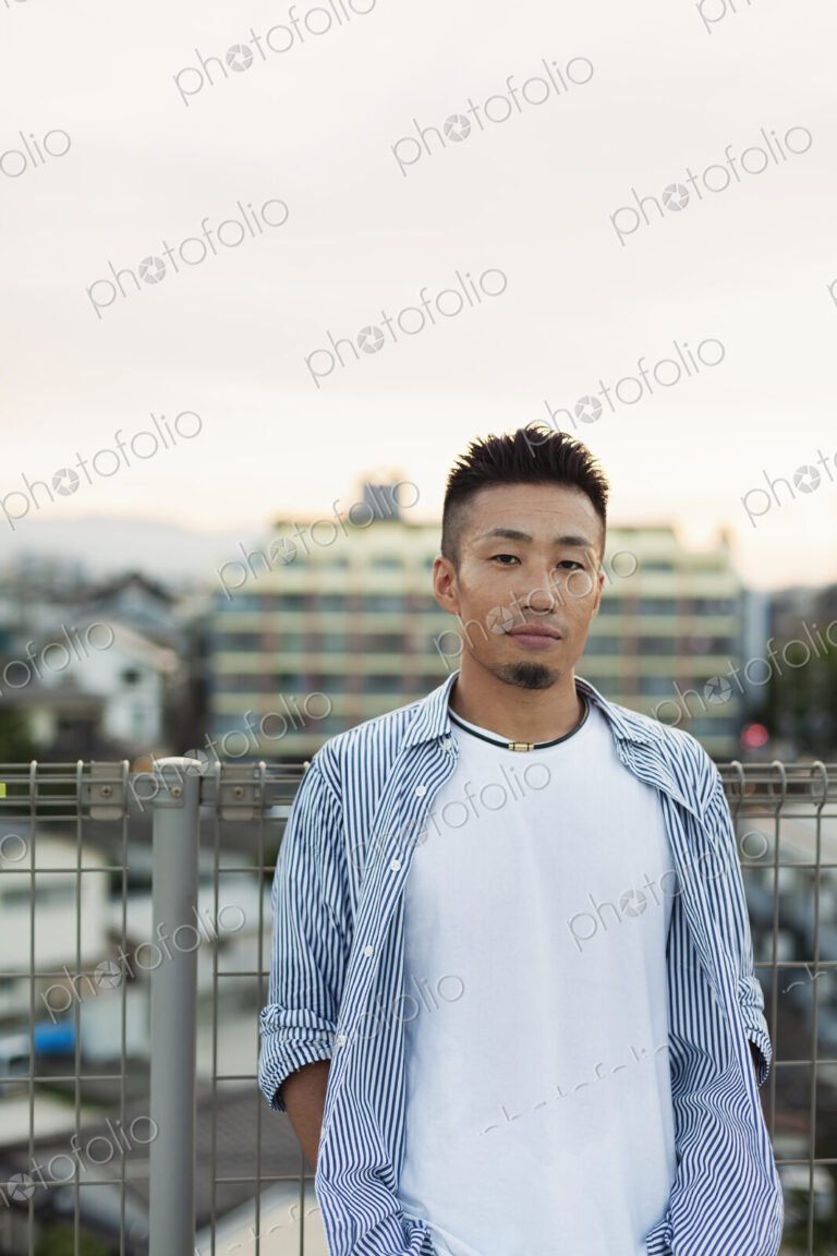 Young Japanese man standing on a rooftop in an urban setting, looking at camera.