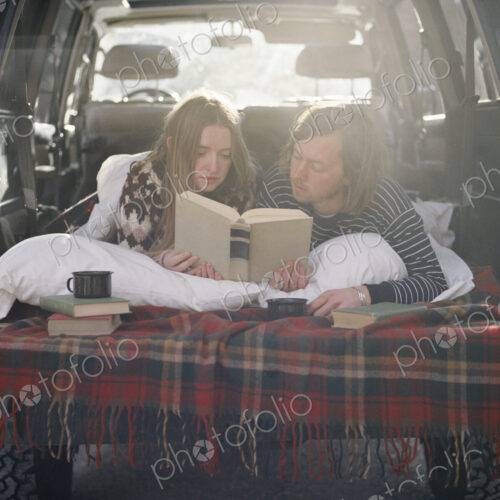 Young couple lying in the back of their car, reading a book.