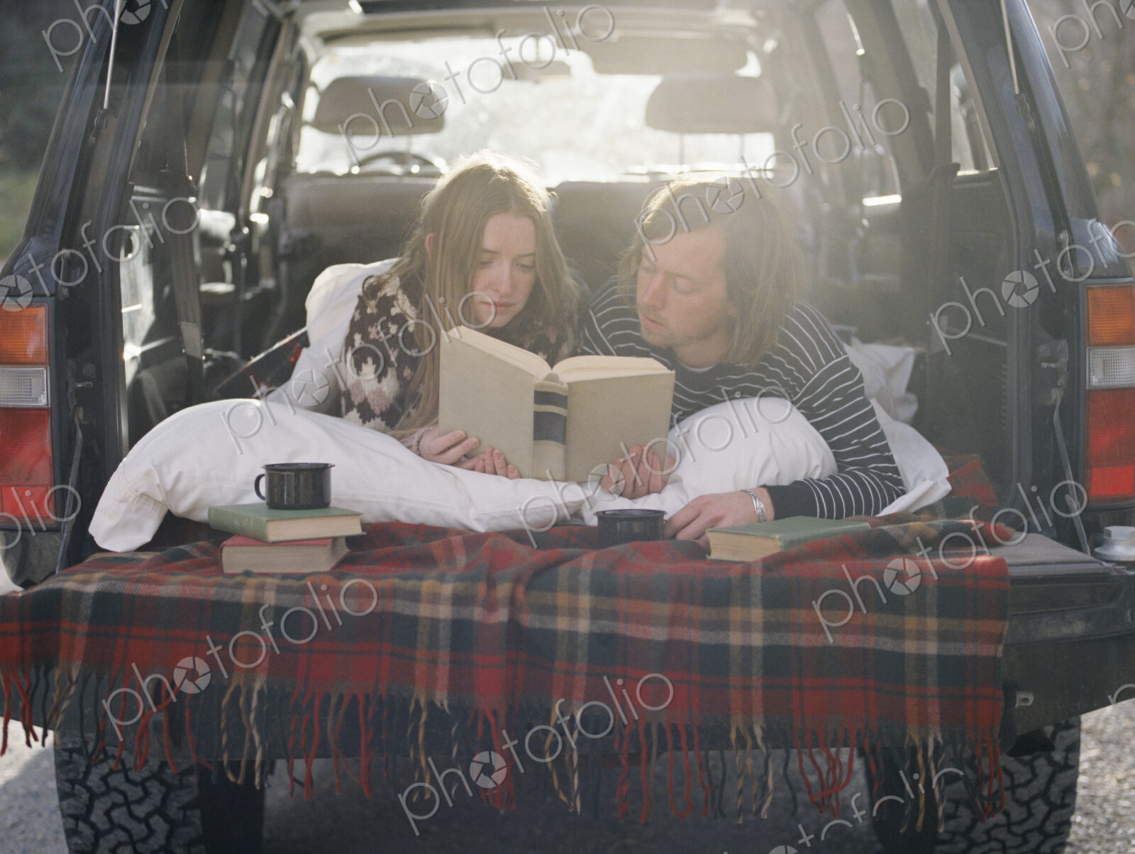 Young couple lying in the back of their car, reading a book.