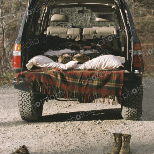 Young couple sleeping in the back of their car, boots standing on the ground in… Young couple sleeping in the back of their car, boots standing on the ground in…