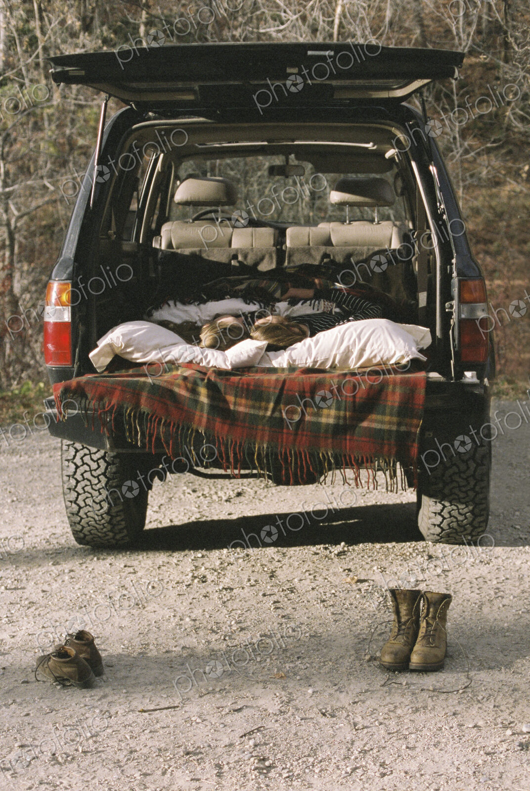 Young couple sleeping in the back of their car, boots standing on the ground in…