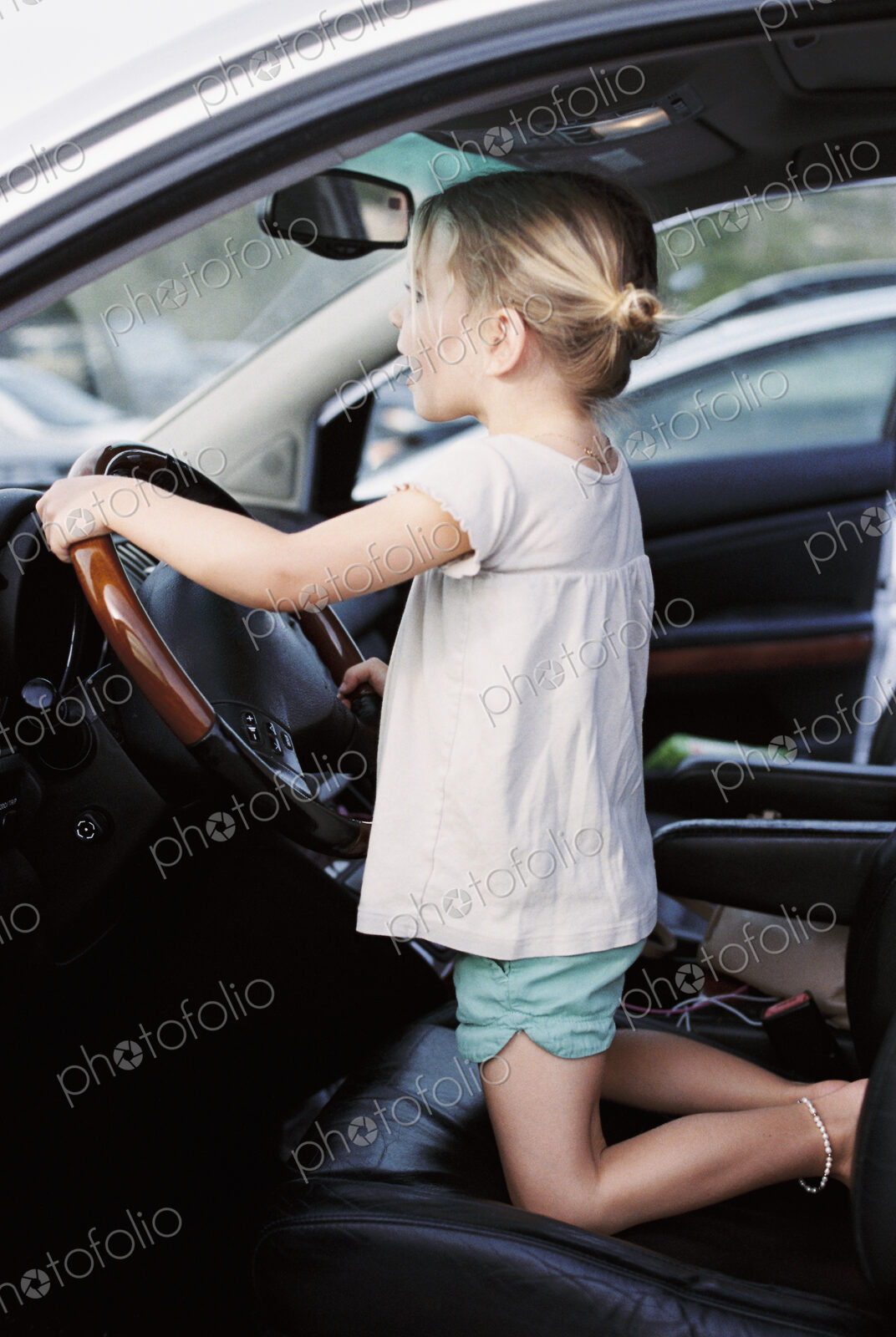 Young girl kneeling on the drivers seat of a car, holding the steering wheel,…