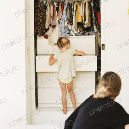 Young girl looking for clothes in a chest of drawers, her mother sitting on a bed,… Young girl looking for clothes in a chest of drawers, her mother sitting on a bed,…