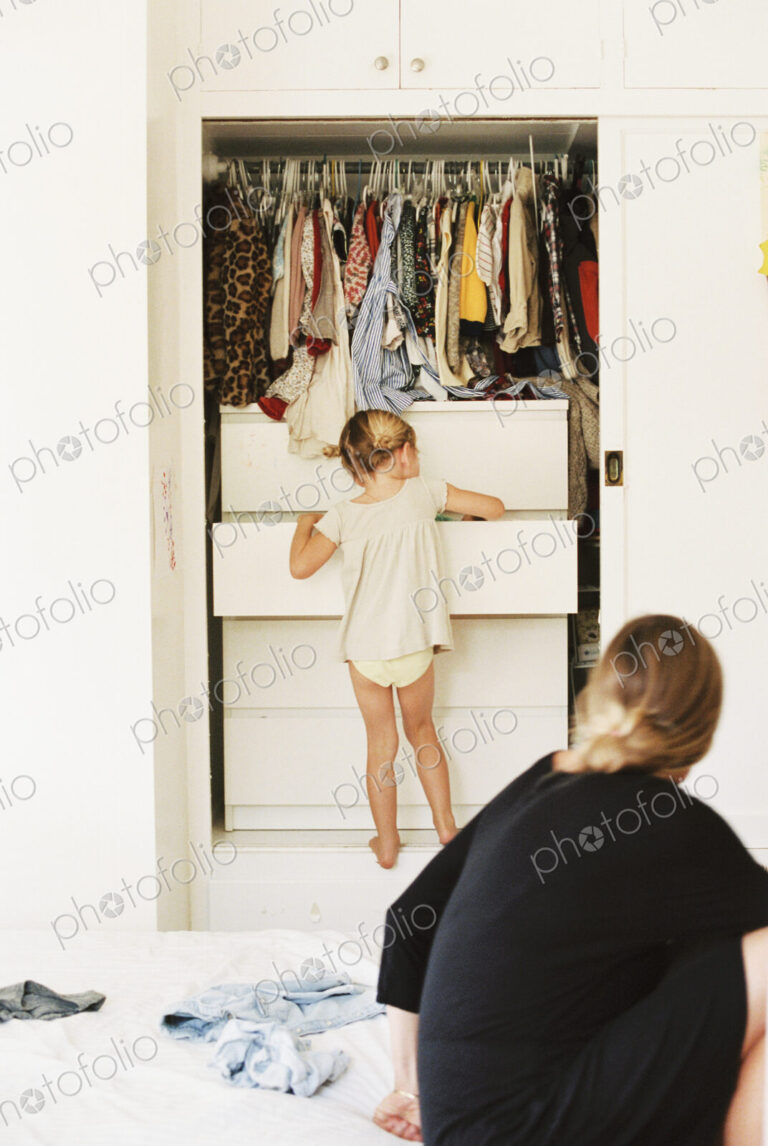 Young girl looking for clothes in a chest of drawers, her mother sitting on a bed,…