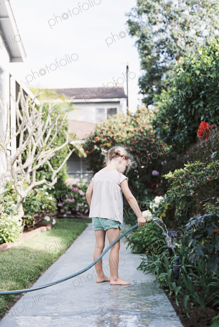 Young girl standing on a path in a garden, playing with a water hose.