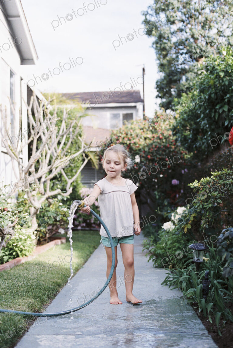 Young girl standing on a path in a garden, playing with a water hose.