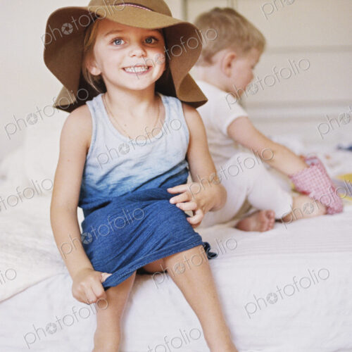 Young girl wearing a dress and large floppy hat on a bed with her younger brother. Young girl wearing a dress and large floppy hat on a bed with her younger brother.