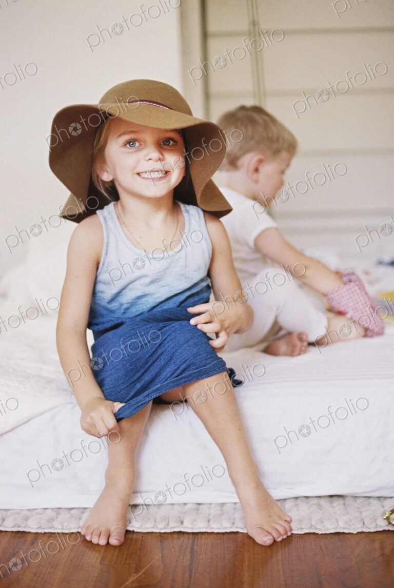 Young girl wearing a dress and large floppy hat on a bed with her younger brother.