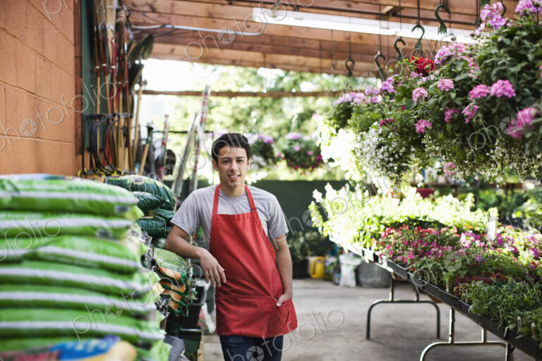 Young man employee in a red apron leaning against bags of compost at a garden center…