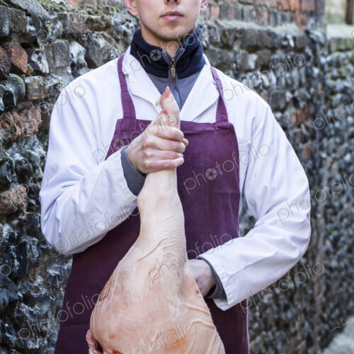Young man wearing apron standing outdoors, holding ham, looking at camera.