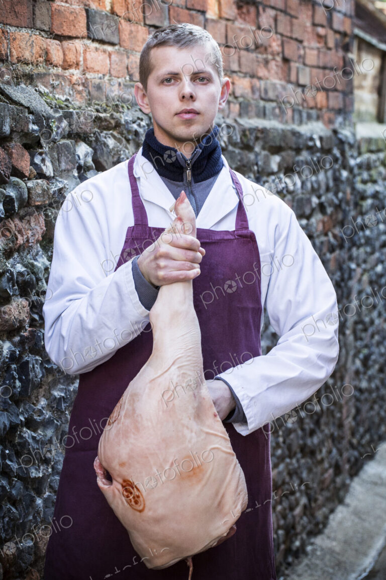 Young man wearing apron standing outdoors, holding ham, looking at camera.
