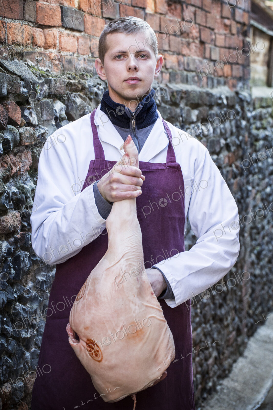 Young man wearing apron standing outdoors, holding ham, looking at camera.